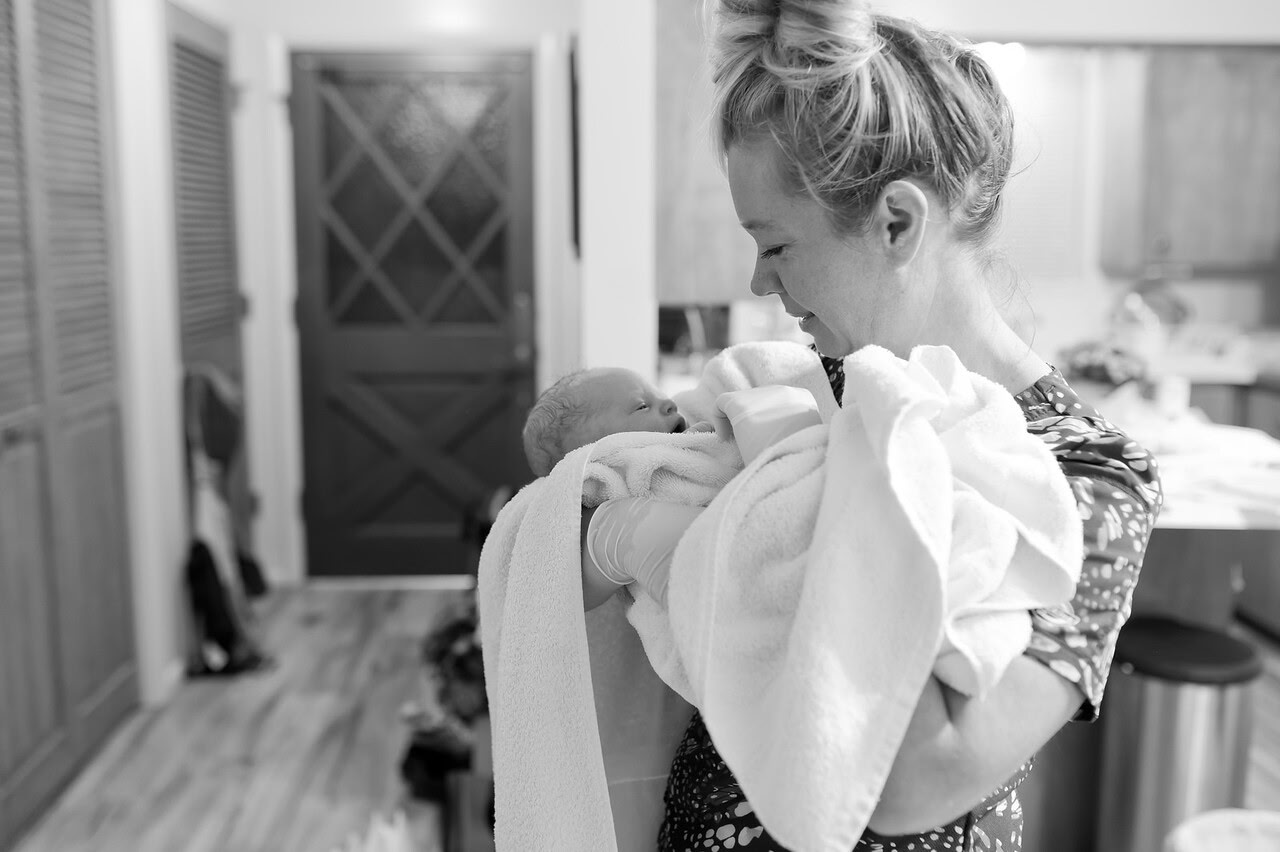 BenNi, wearing gloves, smiles down at a newborn wrapped in a towel in a family's home